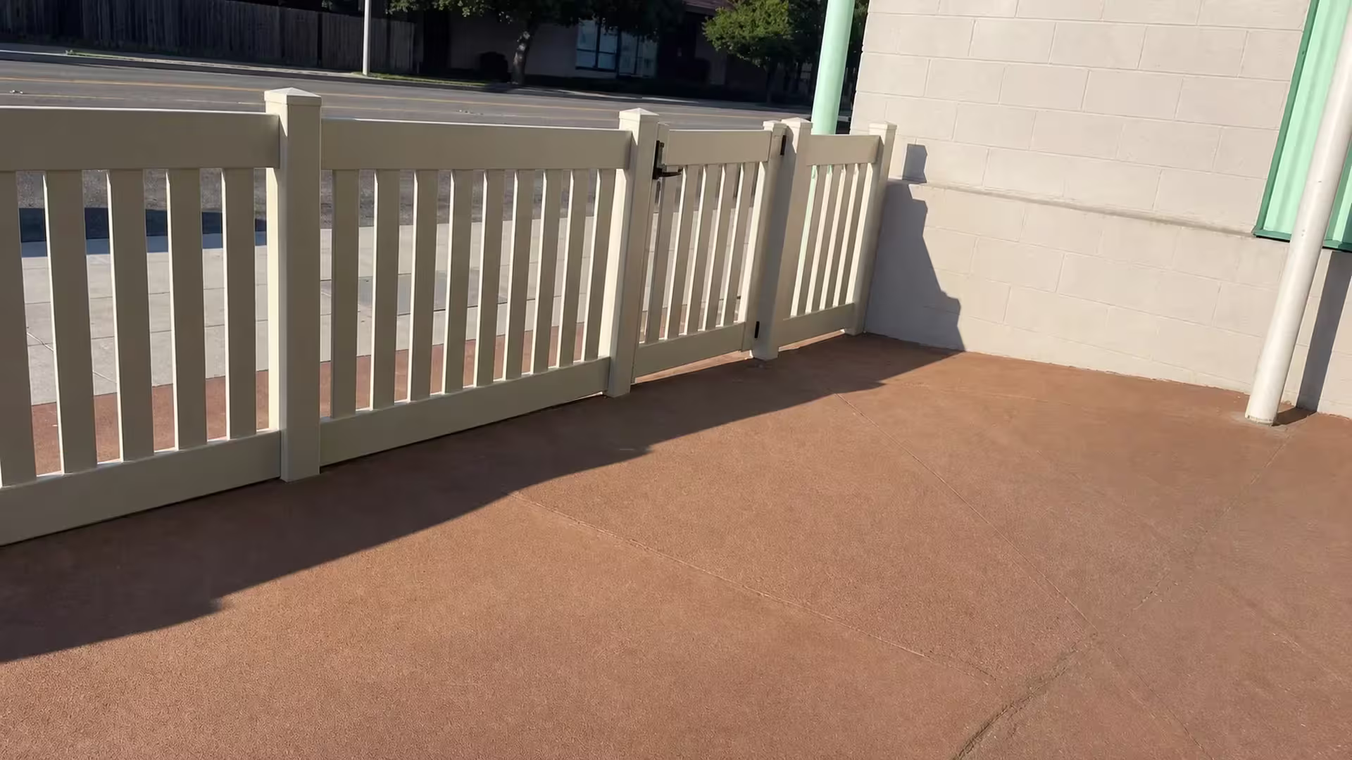 A beige block wall runs beside a Tan vinyl fence of closed-top pickets angling toward a masonry building