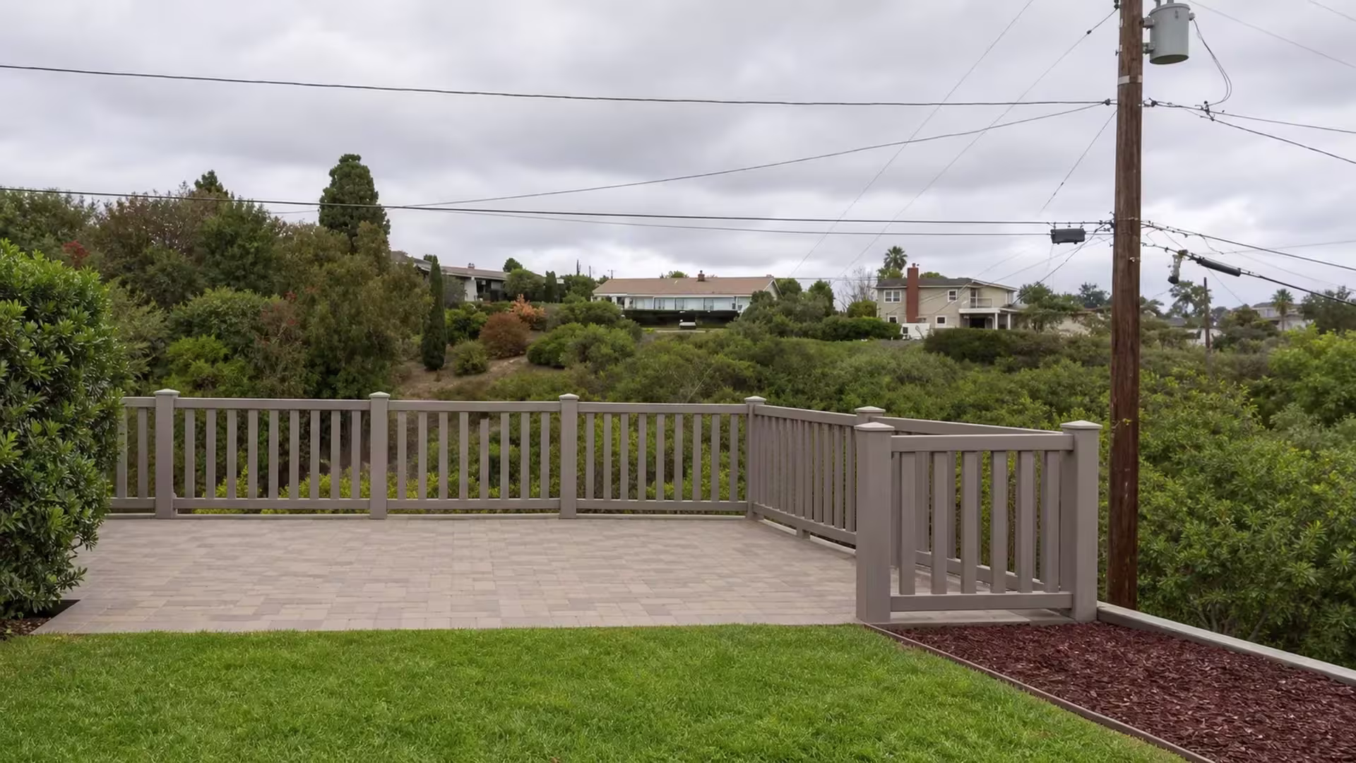 A raised patio slab is ringed by a Weathered Blend closed-top picket fence in vinyl above a mulch bed