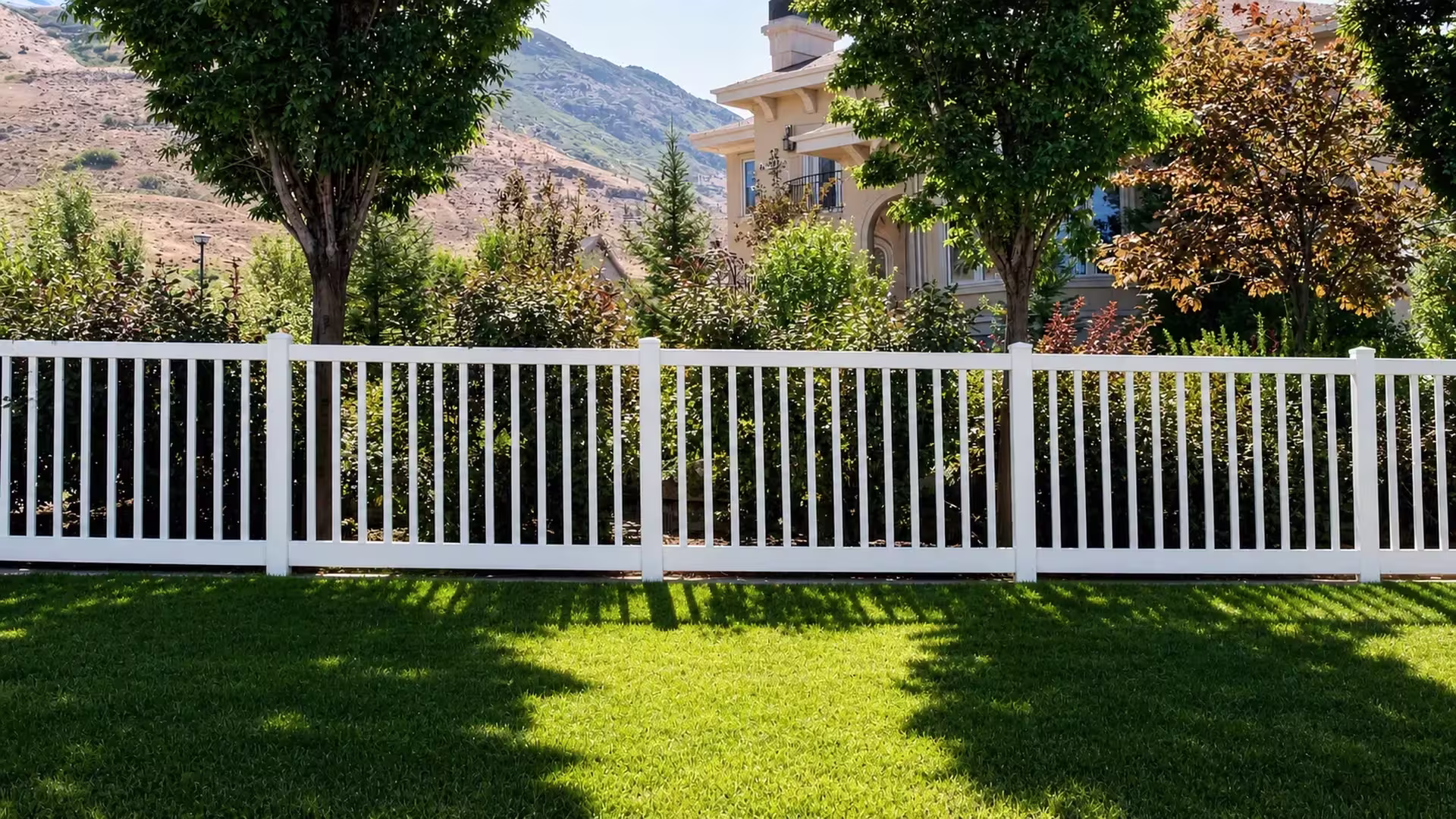 Mature trees and a mountain backdrop rise behind a level White vinyl closed-top picket fence crossing the lawn
