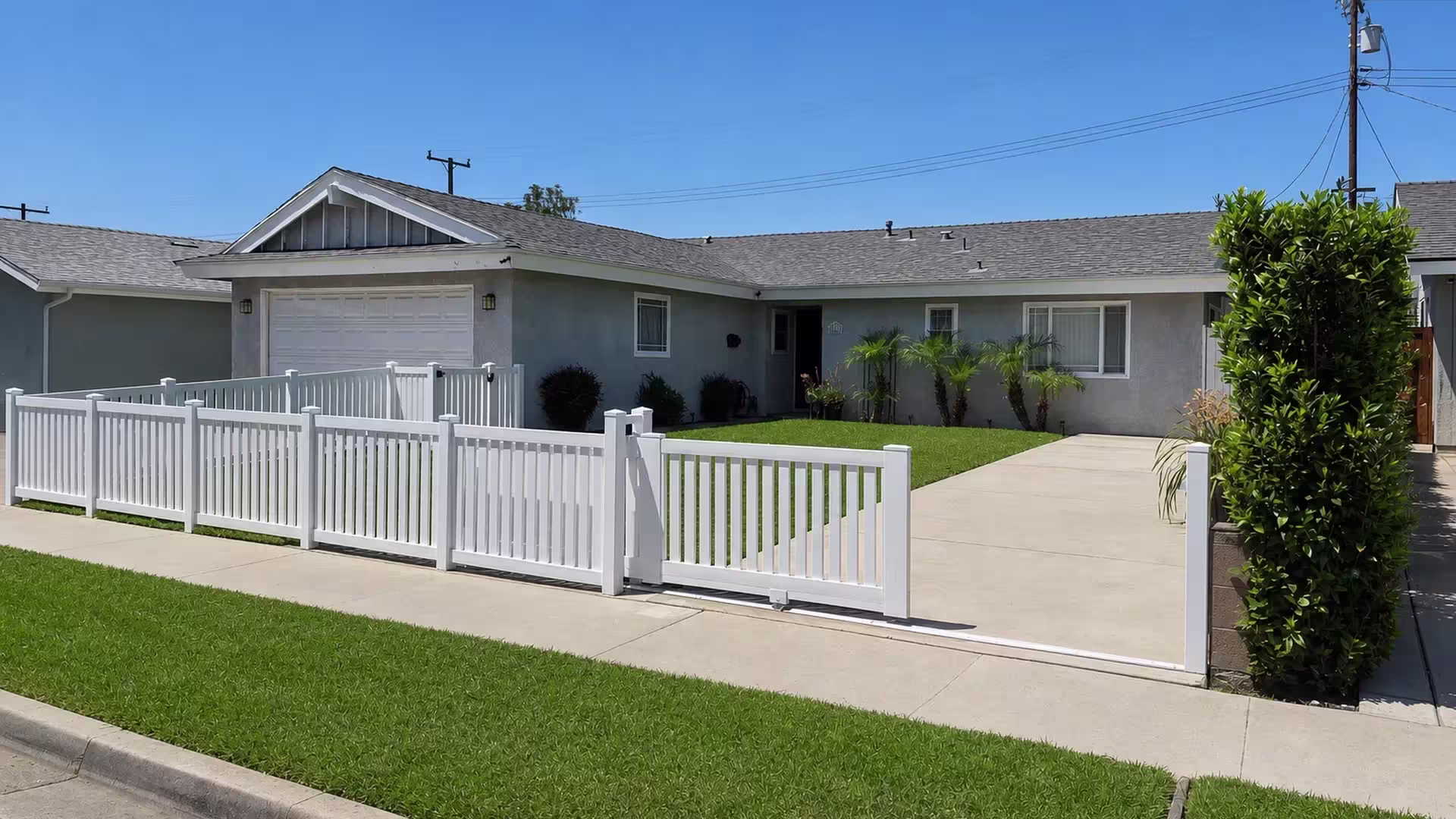 Past a gray house and garage, this White vinyl picket fence ends at a sliding driveway gate with its roller track