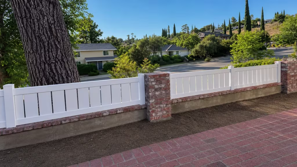 A short stretch of White vinyl picket fence tops a low masonry wall, ending at a red-and-gray brick pier