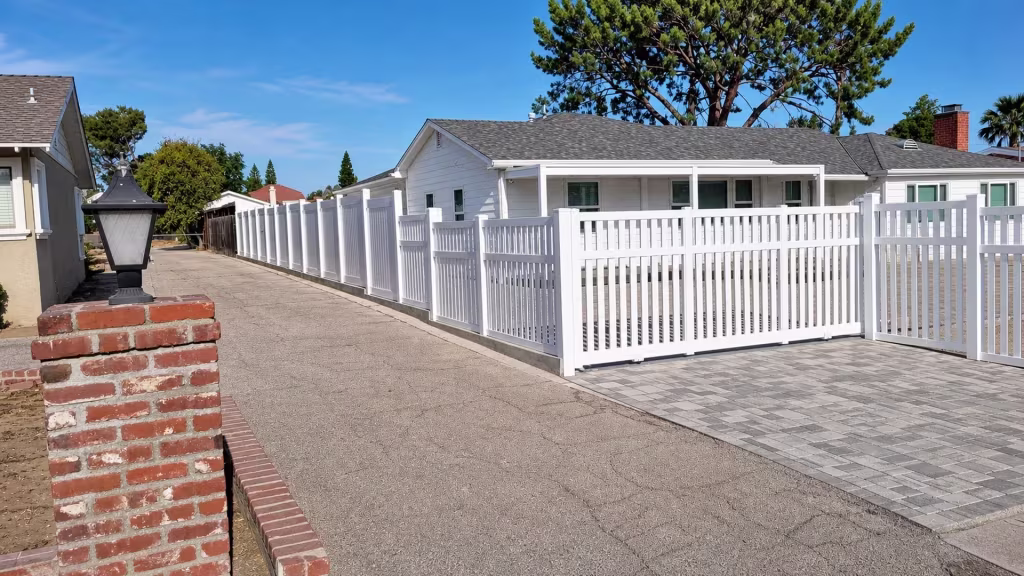 Two gate leaves swing open where this White vinyl picket fence meets a brick-and-lantern pillar at the driveway