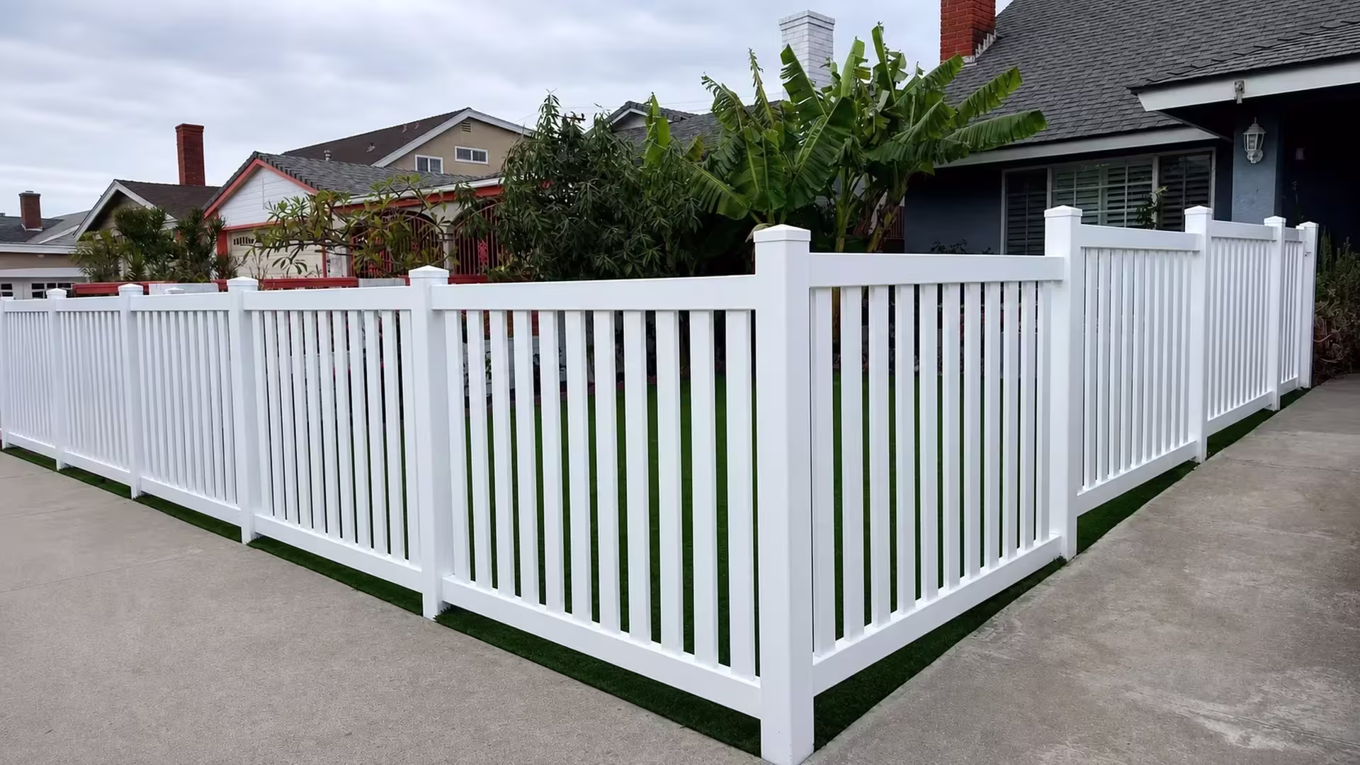 Banana plants line the corner where a White vinyl picket fence turns toward a blue-gray house