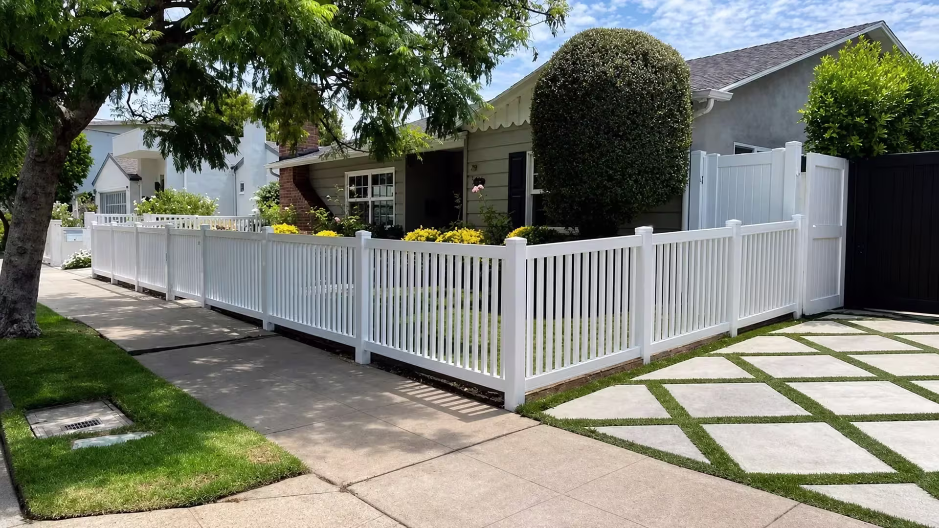 A sculpted hedge and a grass-jointed driveway border this White vinyl picket fence at its L-shaped corner