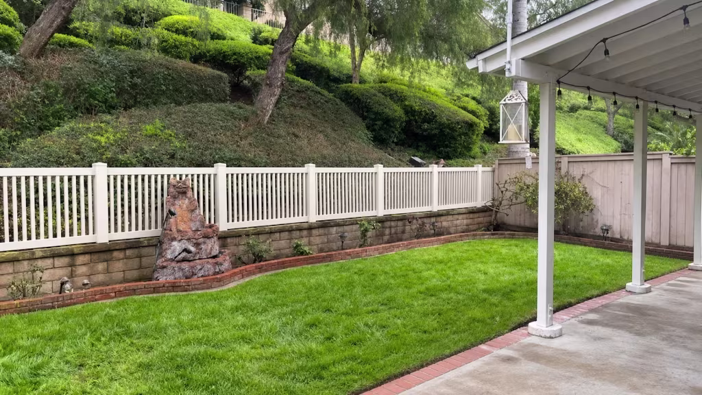 A rock water feature and hanging lantern sit near a White vinyl picket fence that caps a curved wall