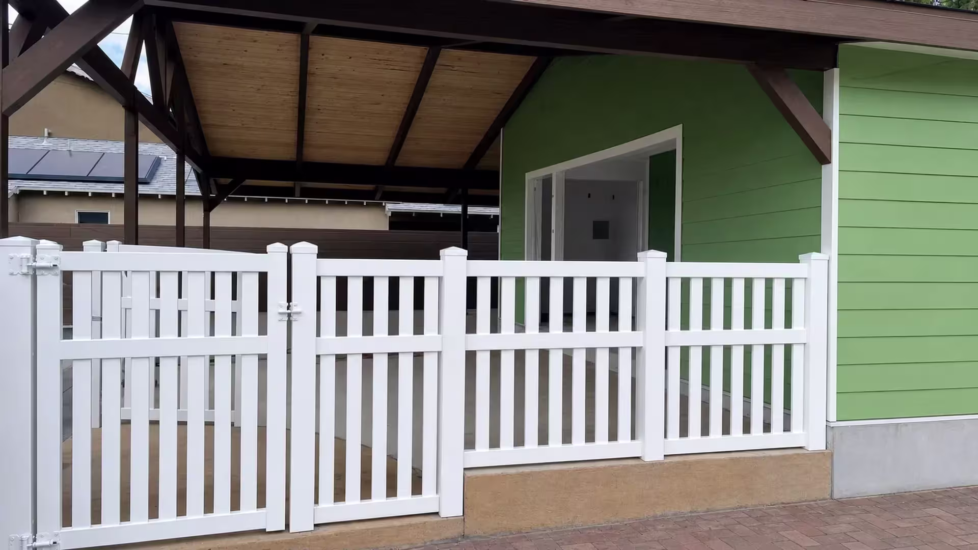 An eight-picket gate leaf on a White vinyl fence, latched at the right, opens beside a covered patio