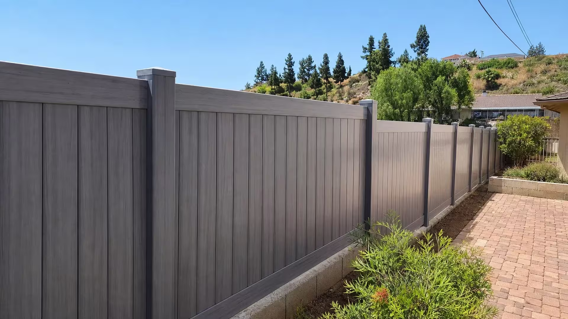 A low concrete curb and brick path border this Coastal Cedar vinyl privacy fence along the hillside