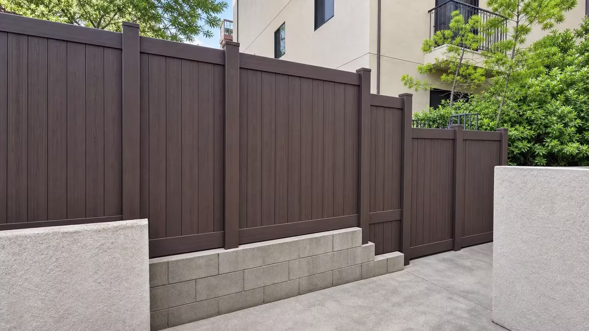 Balcony railings and a downpipe overlook this Dark Walnut vinyl privacy fence along a sloped concrete path