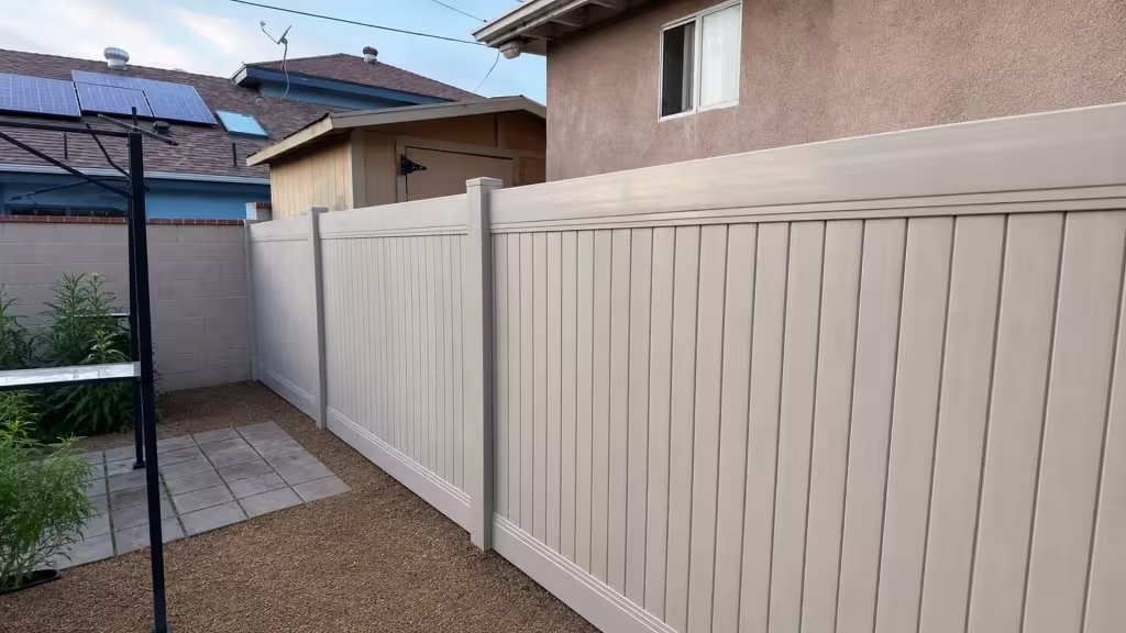 Tan vinyl vertical privacy fence edges a pale masonry boundary near a stucco outbuilding