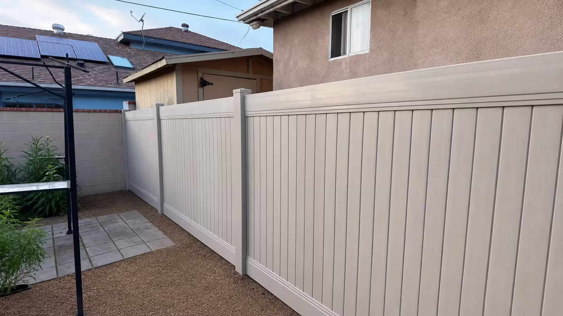 Tan vinyl vertical privacy fence edges a pale masonry boundary near a stucco outbuilding