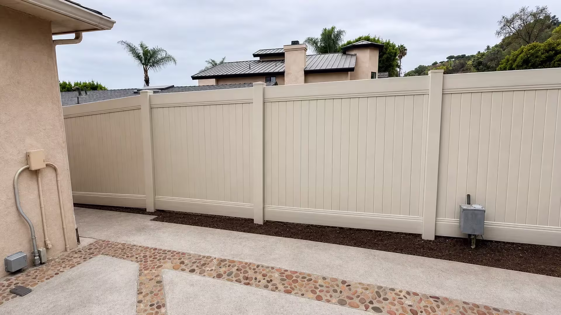 Pebble banded patio paving borders a Tan vinyl vertical privacy fence below a distant hillside