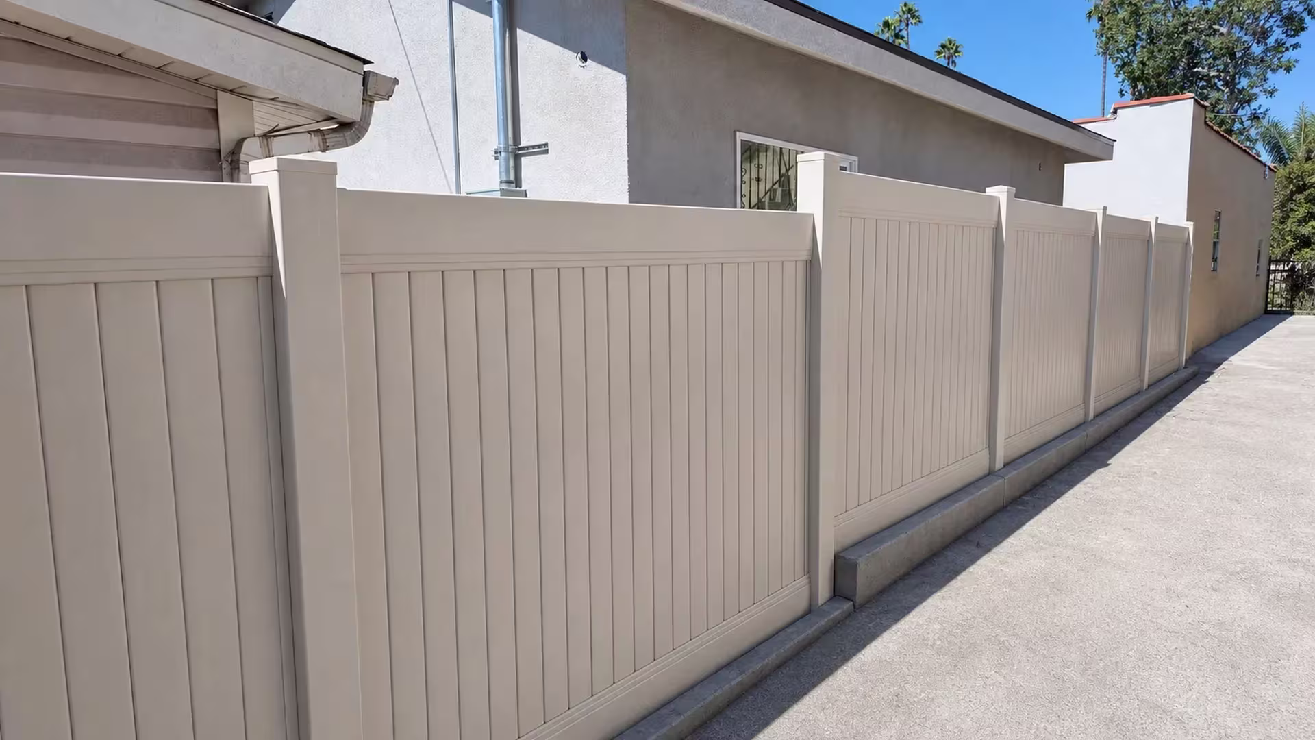 Barred window and concrete curb sit beside a Tan vinyl vertical privacy fence with matching panel construction