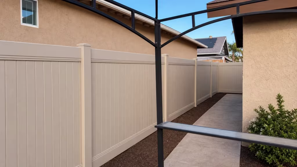 A metal patio cover with curved braces shelters a passage beside a Tan vinyl vertical privacy fence