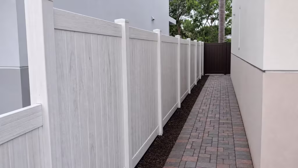 Weathered Aspen vinyl privacy fence lines a narrow side yard beside a red and gray paver walkway