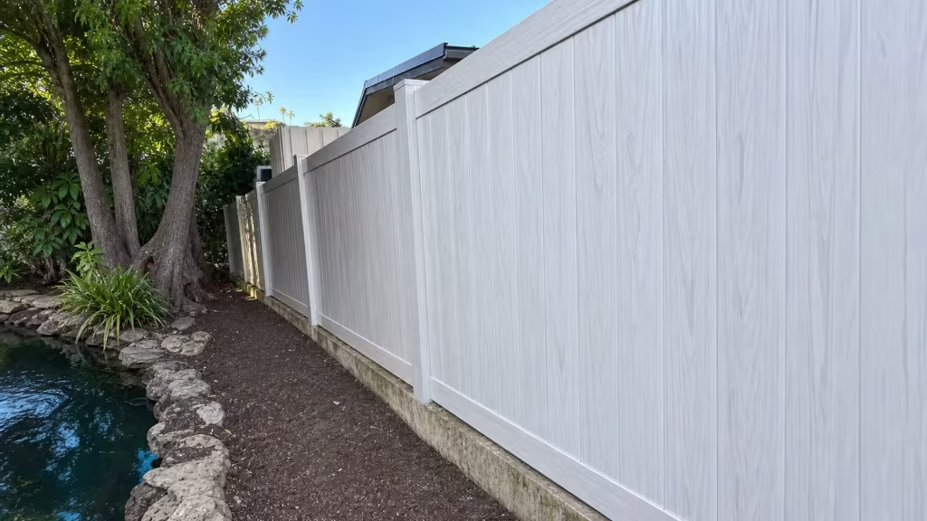 Weathered Aspen vinyl privacy fence sits above a concrete curb along a narrow water edge and rocky soil