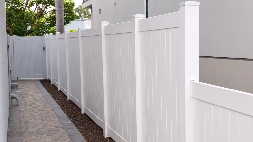 White vinyl vertical privacy fence meets a perpendicular return run beside a narrow soil strip and walkway