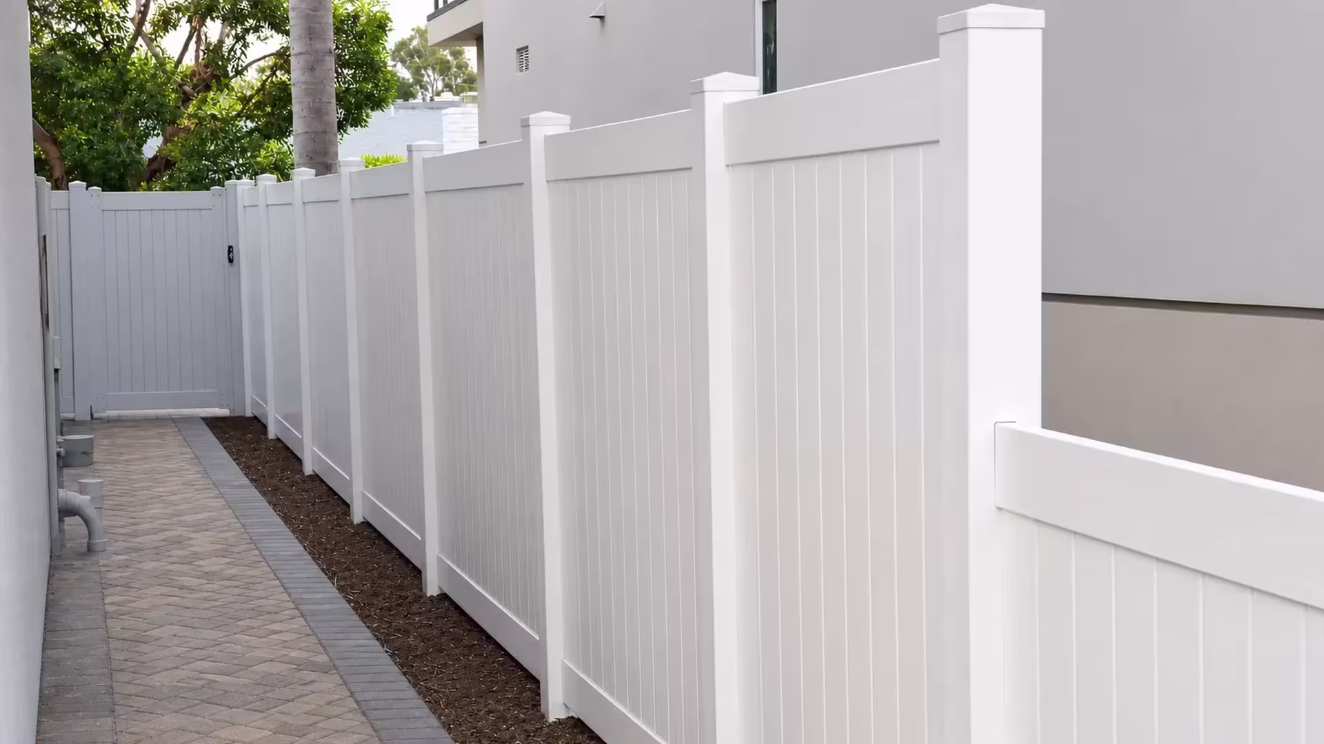 White vinyl vertical privacy fence meets a perpendicular return run beside a narrow soil strip and walkway