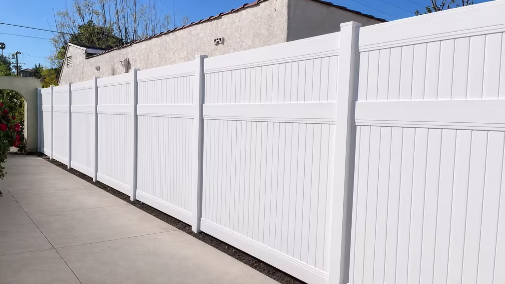 White vinyl vertical privacy fence lines a gray beige stucco outbuilding topped with reddish roof tiles