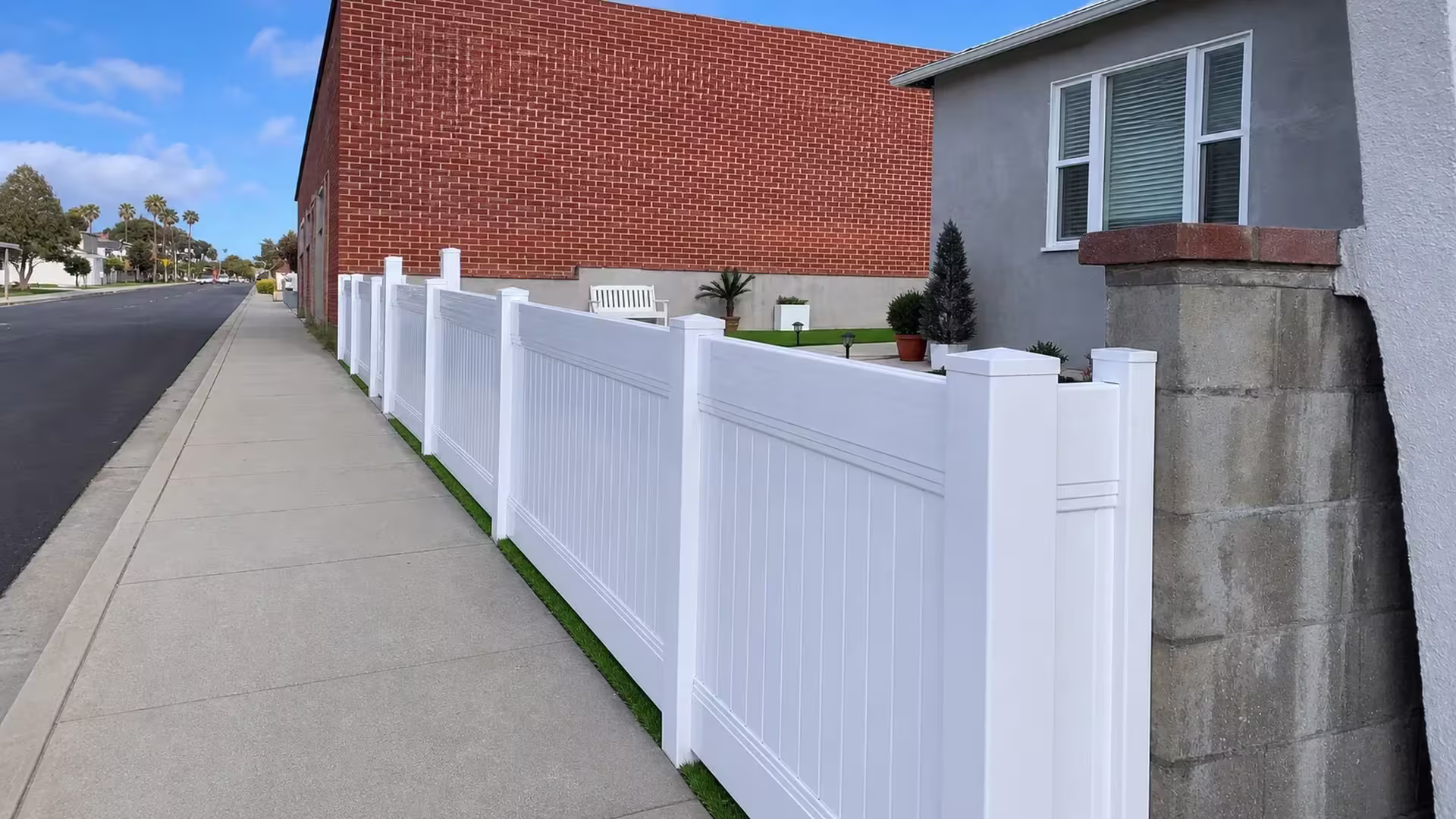 A red brick wall, a bench and the curb border this White vinyl vertical privacy fence as it stretches toward the sidewalk