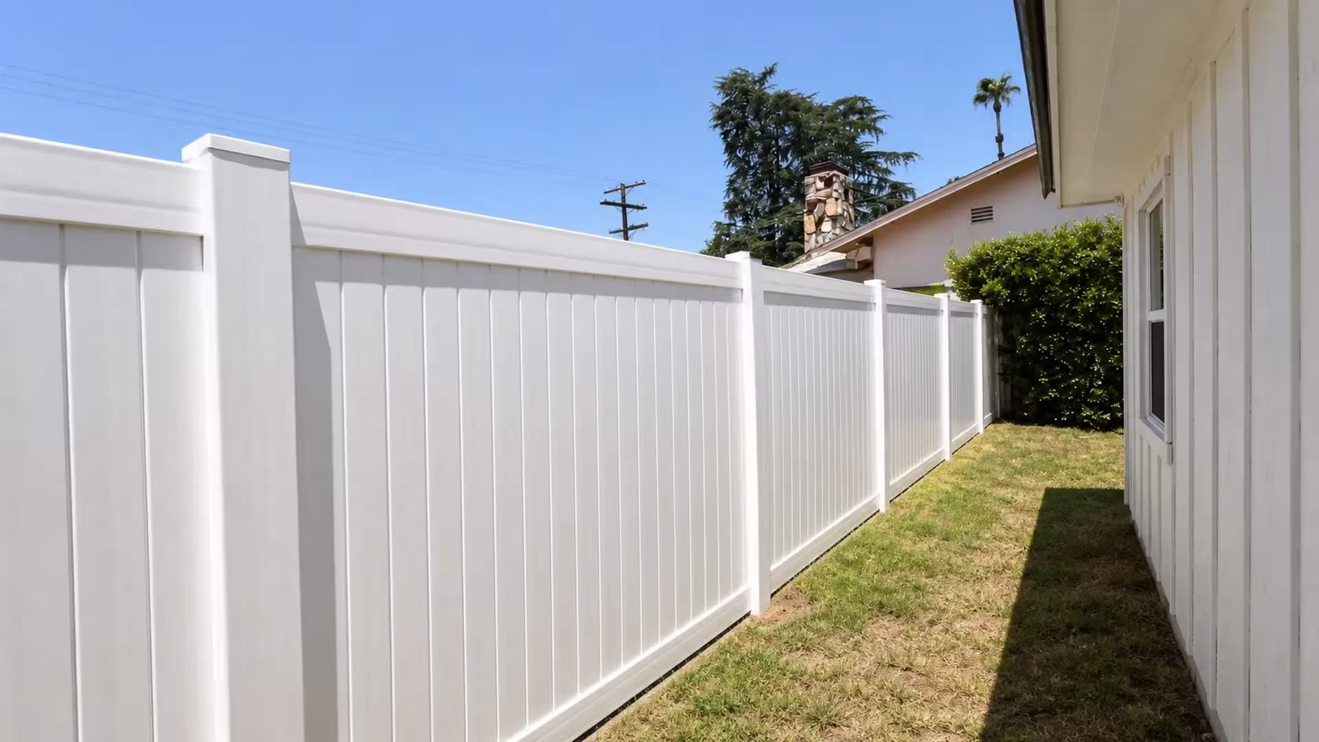 A mature conifer and stone chimney rise beside this White vinyl vertical privacy fence closing the narrow side yard