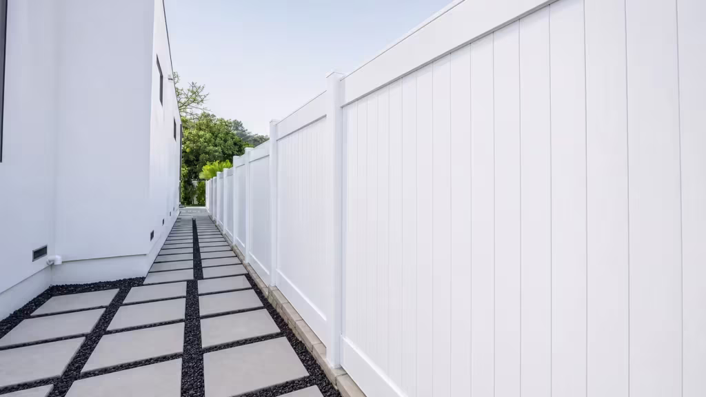 White vinyl privacy fence steps down along concrete slabs with black pebble joints in a narrow passage