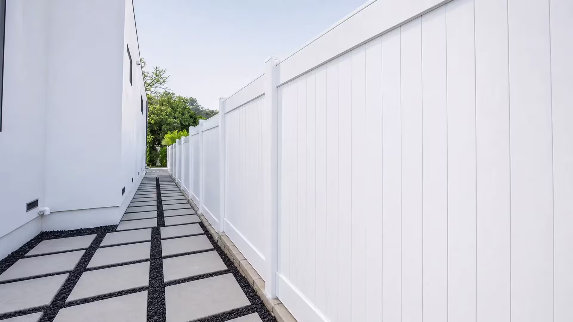 White vinyl privacy fence steps down along concrete slabs with black pebble joints in a narrow passage