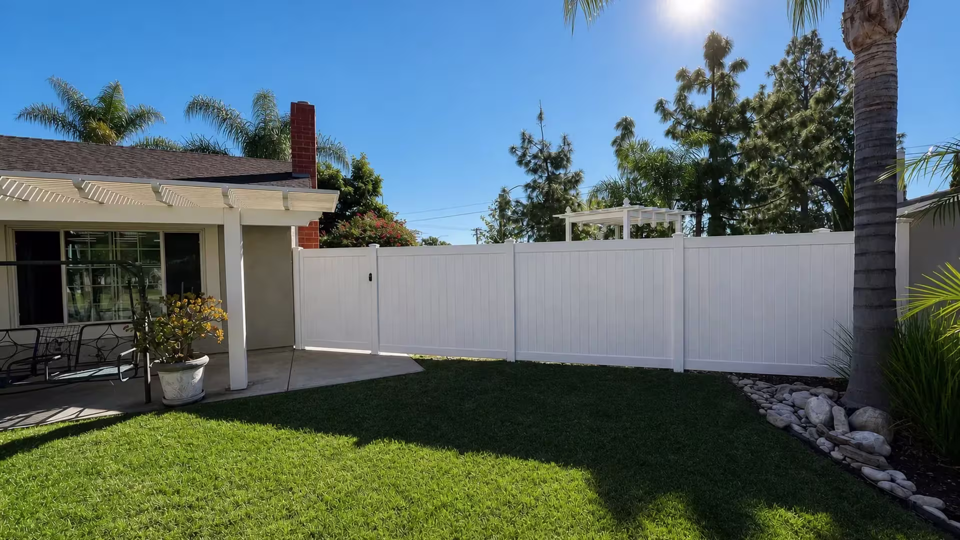 A single gate leaf in this White vinyl vertical privacy fence opens beside a cream stucco house and covered patio