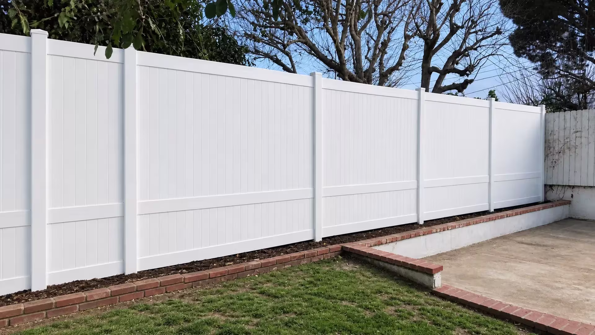 Brick edged walls and an old wood fence flank this White vinyl vertical privacy fence crossing a raised planting strip