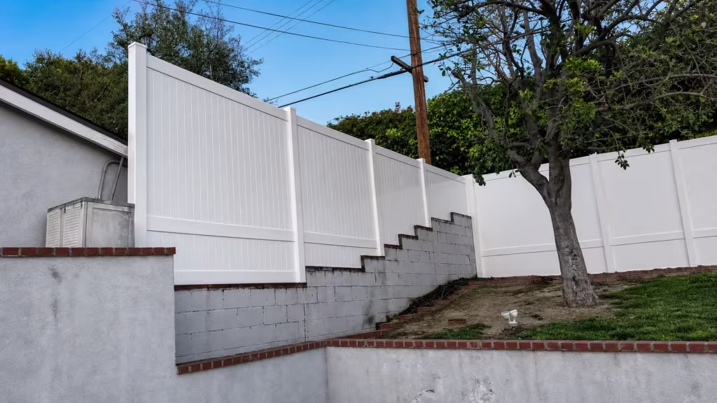 White vinyl vertical privacy fence follows a sloped residential yard above painted masonry retaining walls and steps