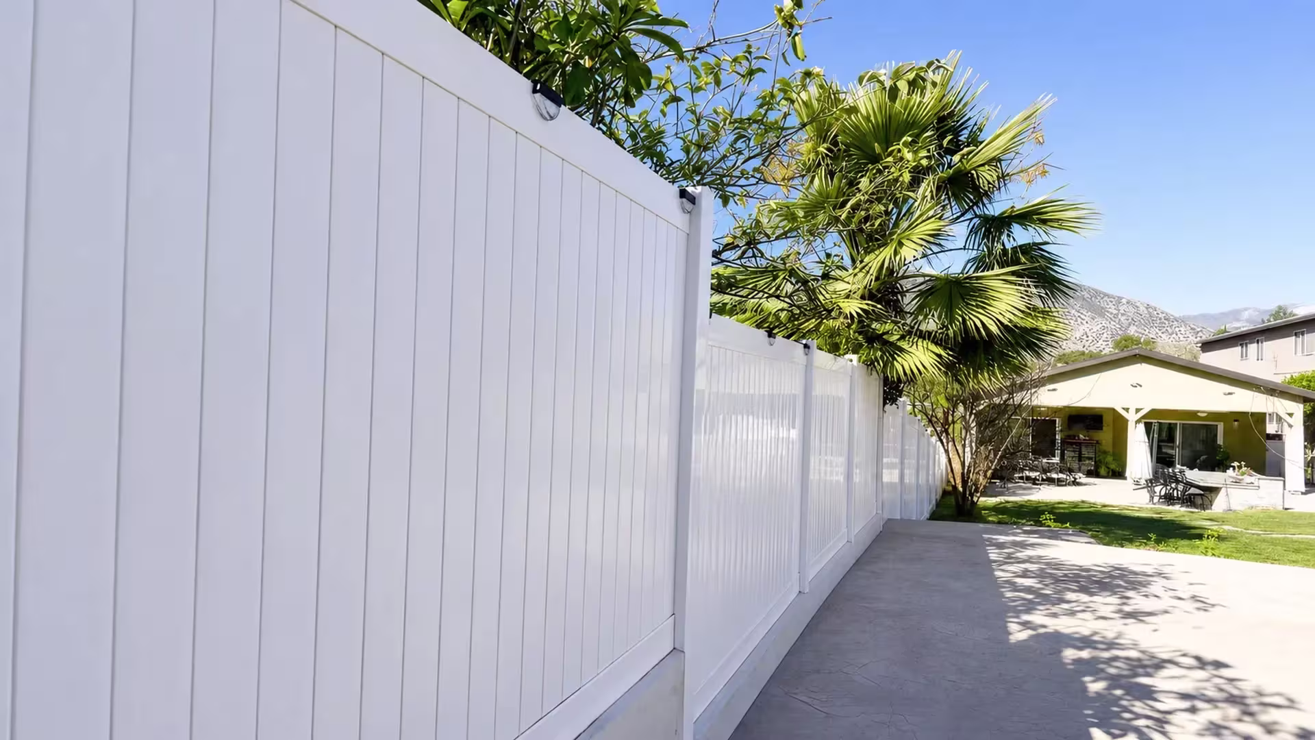White vinyl privacy fence stretches across stamped concrete paving toward a backyard patio and distant mountains