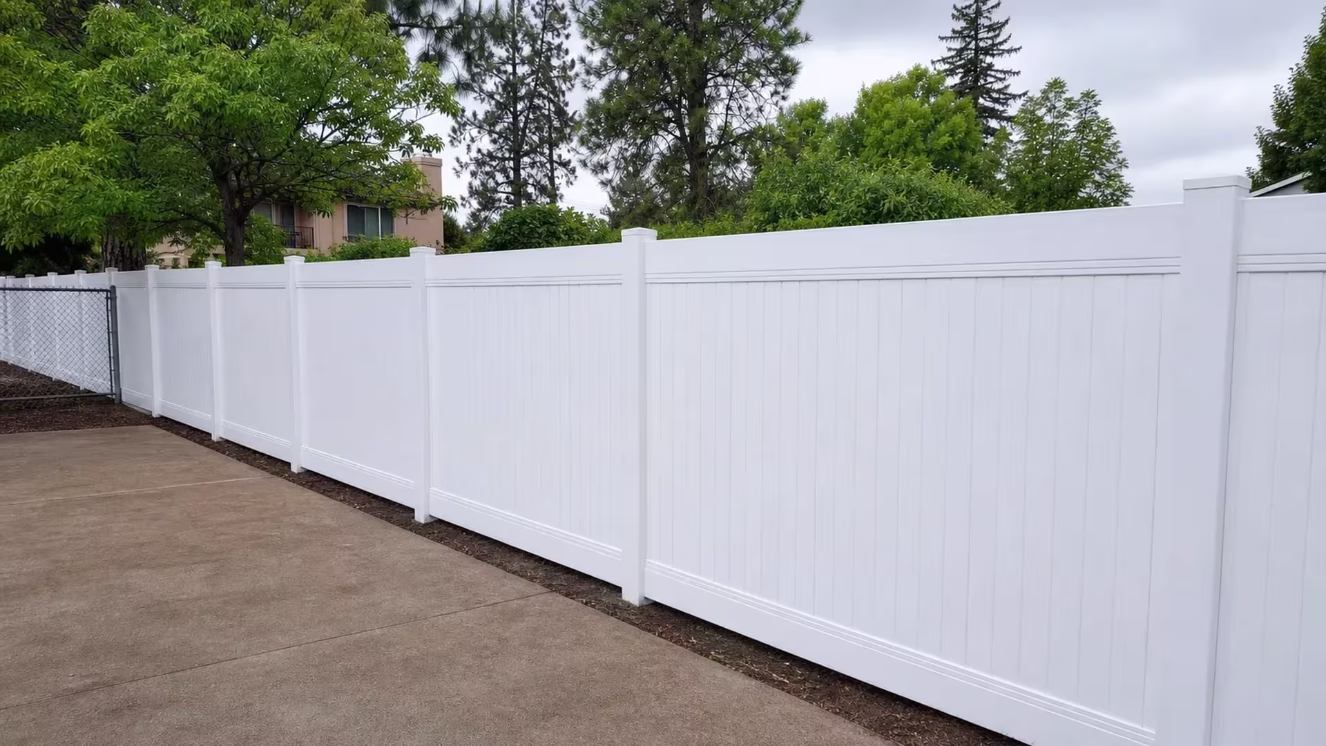 Five panel spans of White vinyl vertical privacy fence step gently downhill past a house on sloped ground