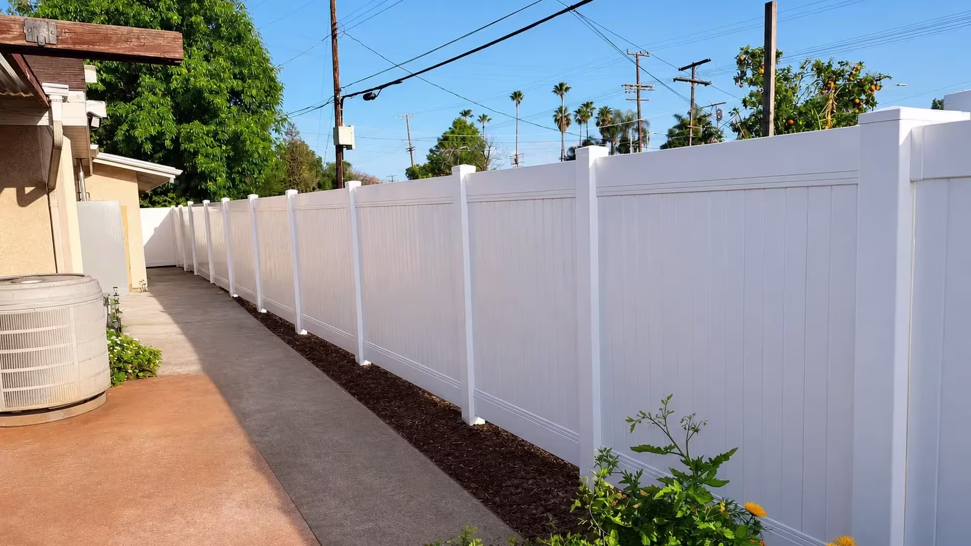 Outdoor condenser and reddish walking surfaces line this White vinyl vertical privacy fence along the side yard