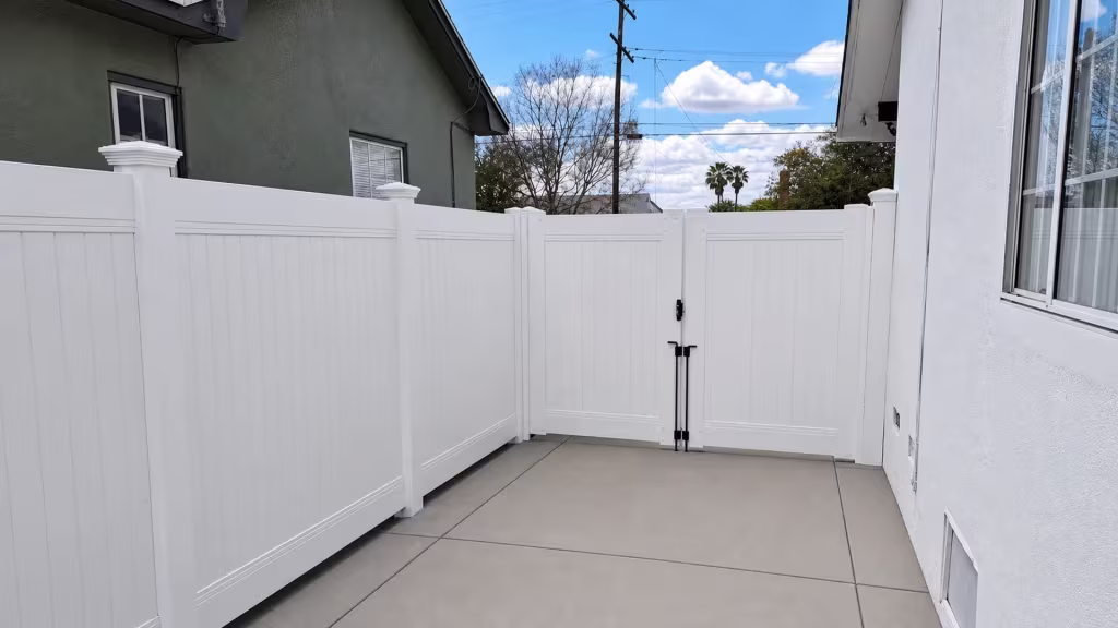 A jointed concrete passage runs beside this White vinyl vertical privacy fence before it turns into a two leaf gate