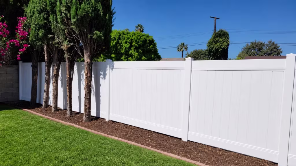 White vinyl privacy fence partly screened by tree trunks beside a curved brick edged flower bed