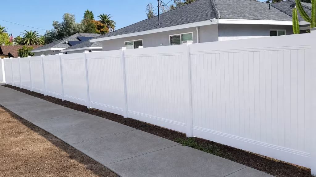 Chain link fencing and a cactus stand beside this White vinyl vertical privacy fence near the concrete sidewalk