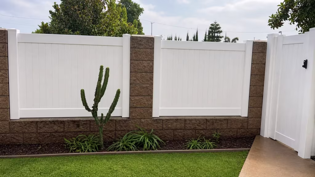 Paired White vinyl vertical privacy fence divides at a central pier over a brown split-face masonry wall