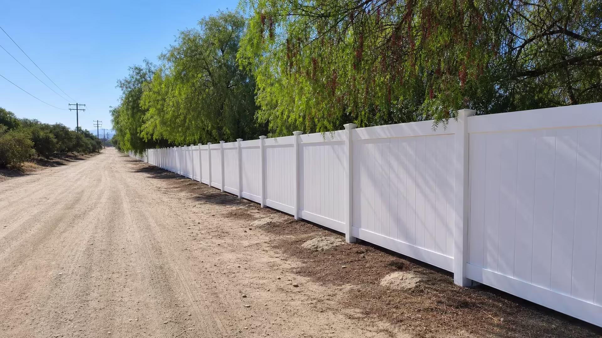 Continuous White vinyl vertical privacy fence extends beside an unpaved rural road toward the horizon