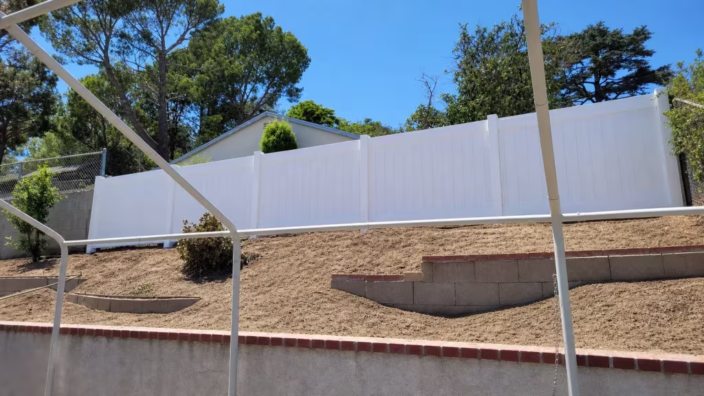 Uniform White vinyl vertical privacy fence descends in stepped panels down a terraced slope beneath a patio cover