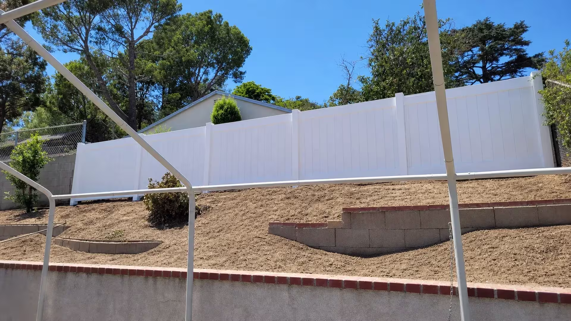 Uniform White vinyl vertical privacy fence descends in stepped panels down a terraced slope beneath a patio cover