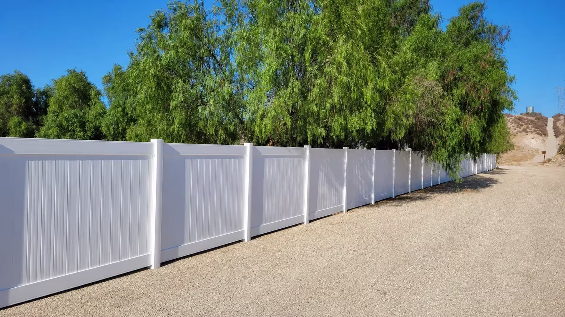 New White vinyl vertical privacy fence continues in a straight run near mature shade trees
