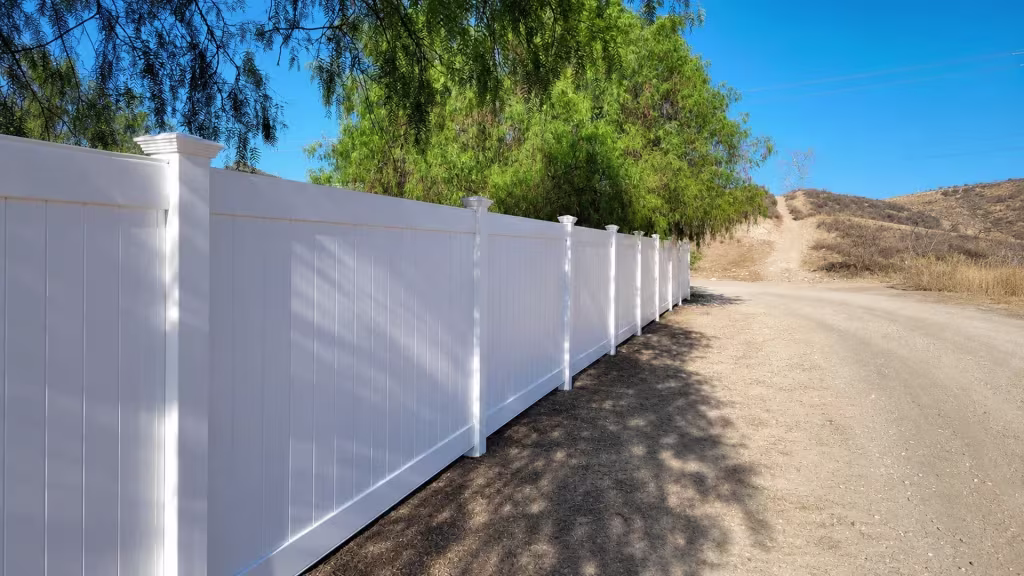 Sturdy White vinyl vertical privacy fence parallels an unpaved hillside access road across dry terrain