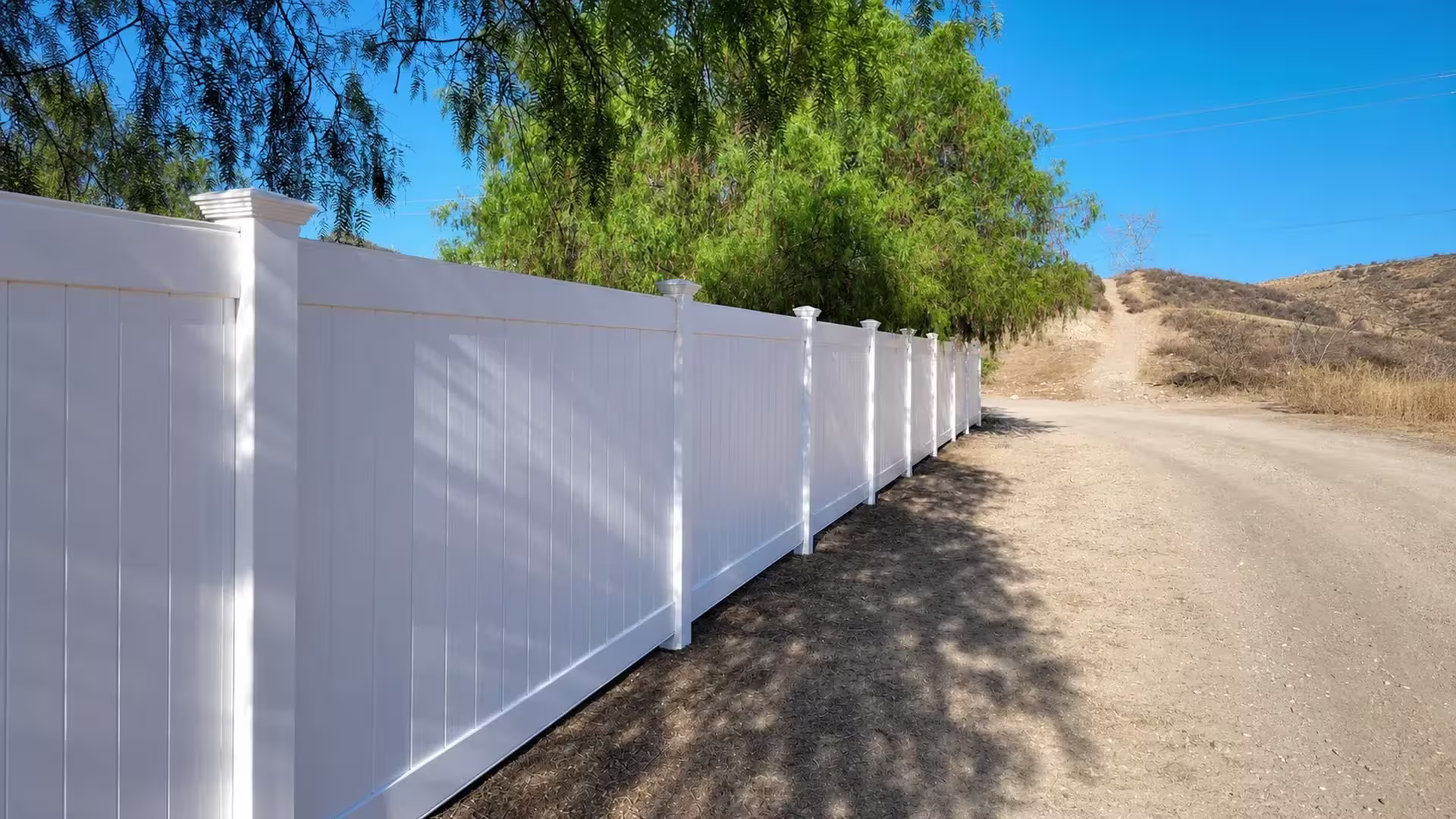 Sturdy White vinyl vertical privacy fence parallels an unpaved hillside access road across dry terrain