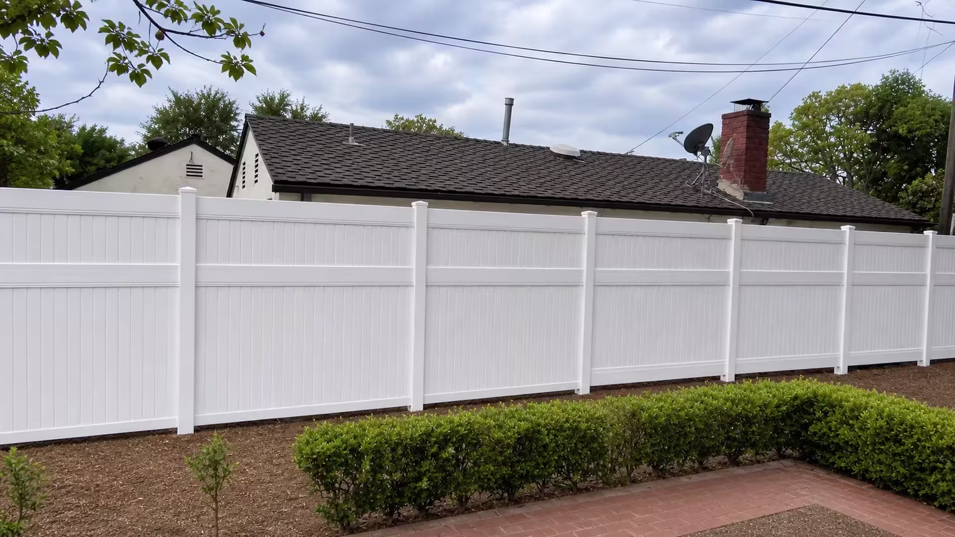 Neat White vinyl vertical privacy fence fronts a brick walkway and trimmed hedge below a gabled roofline