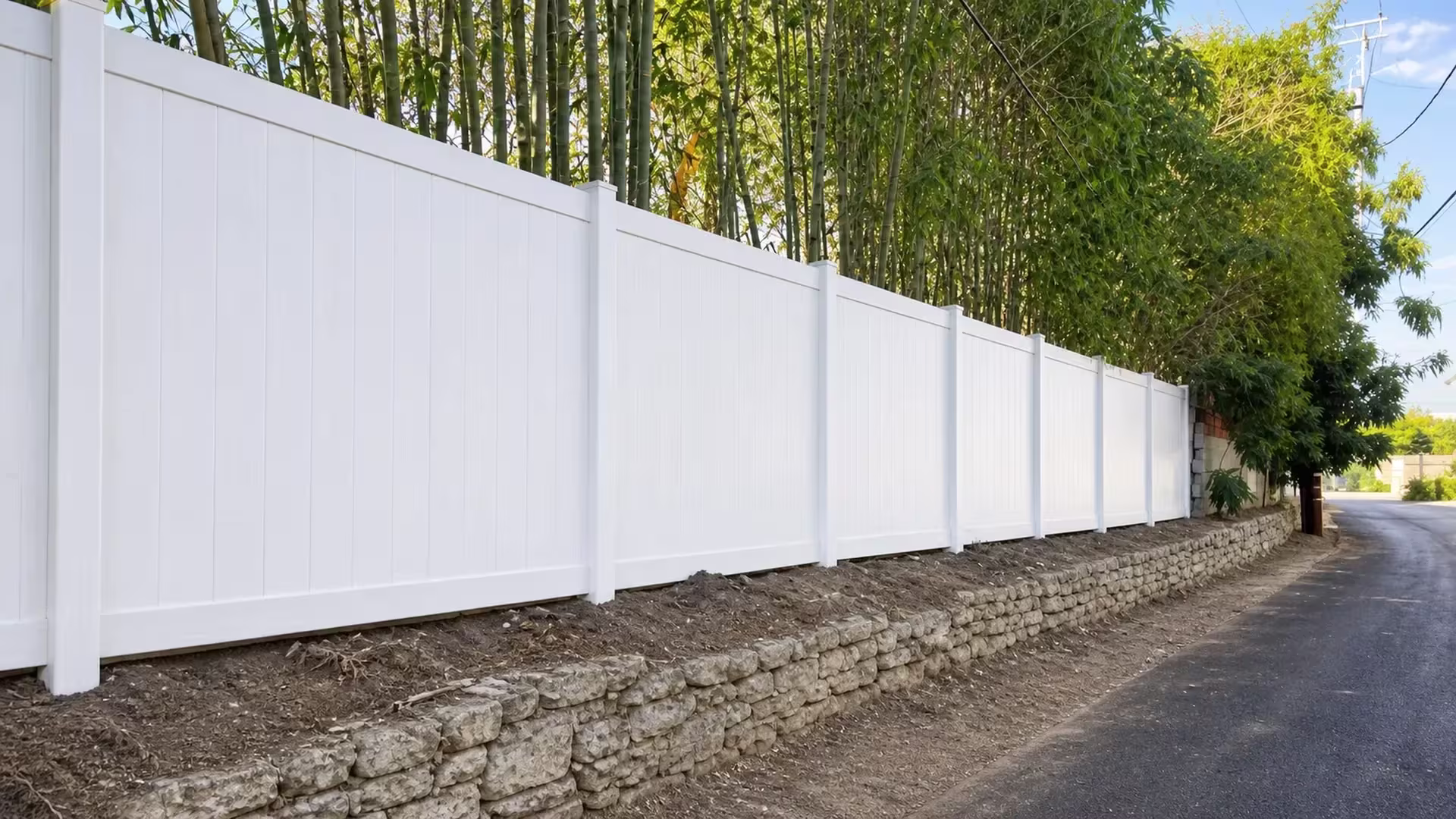 Rigid White vinyl vertical privacy fence hugs an asphalt lane past stacked stone and dense bamboo