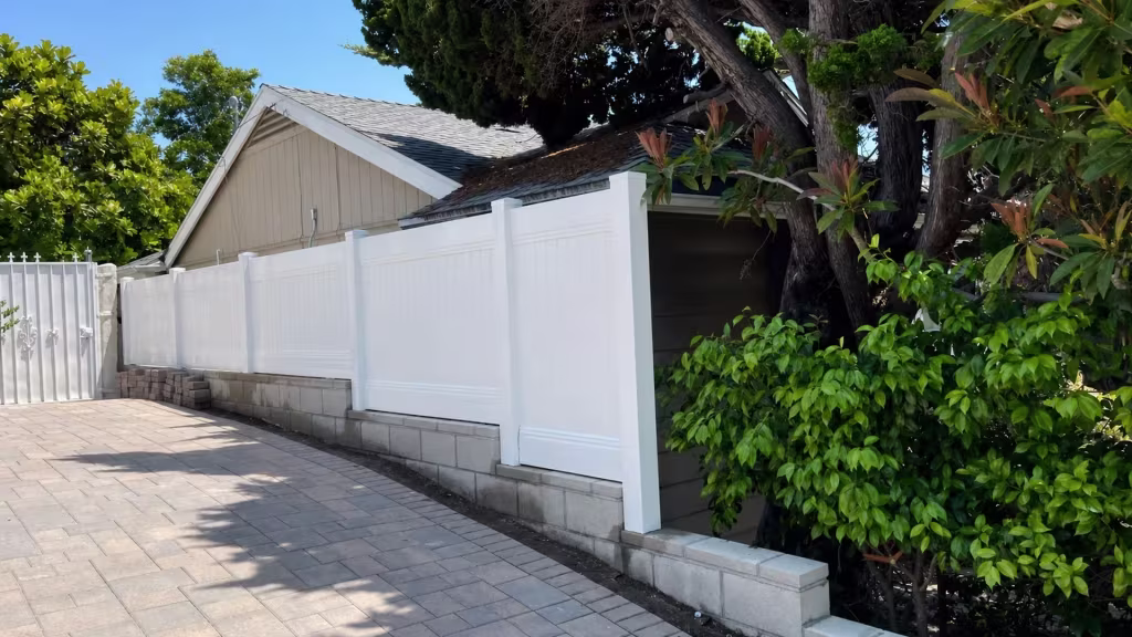 Flush White vinyl vertical privacy fence drops down a gray block retaining wall toward a masonry pier and existing metal gate