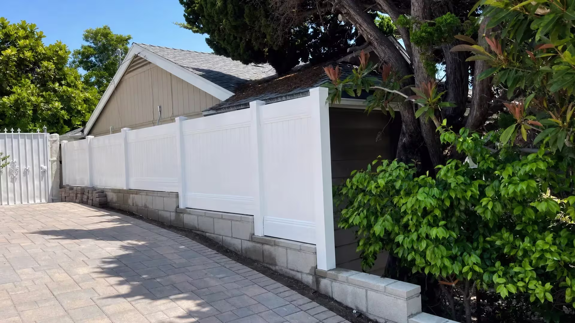 Flush White vinyl vertical privacy fence drops down a gray block retaining wall toward a masonry pier and existing metal gate