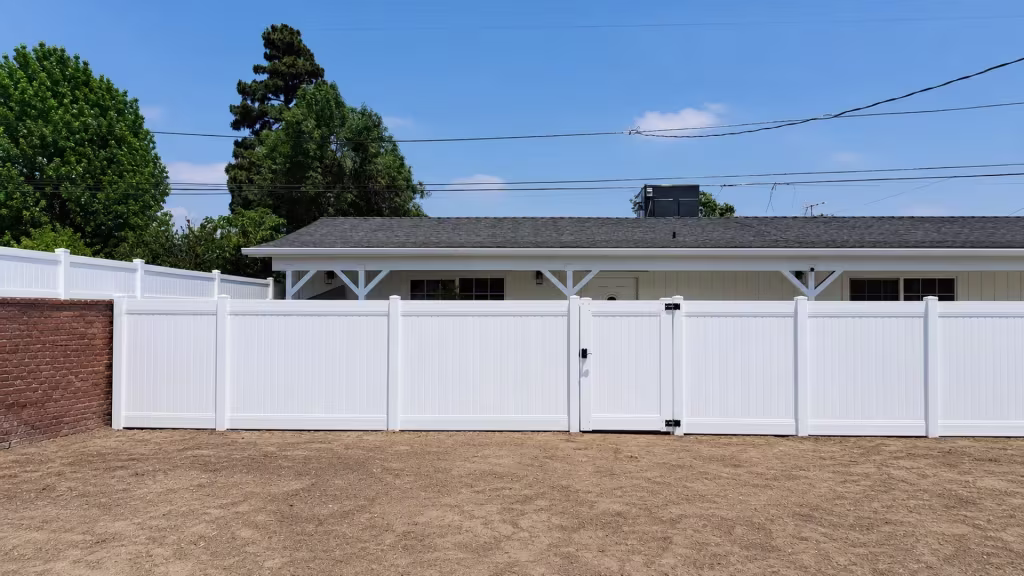 Molded White vinyl vertical privacy fence carries a pedestrian gate with black latch and hinge hardware beside a brick wall