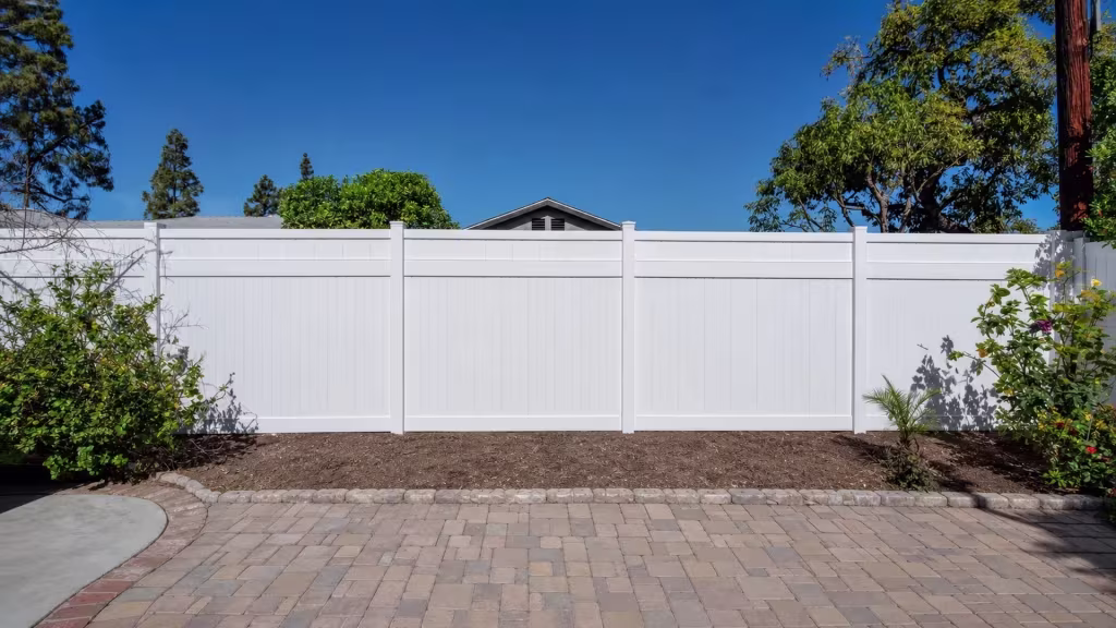 A multicolor paver patio borders this White vinyl privacy fence beside a stone edged raised soil bed