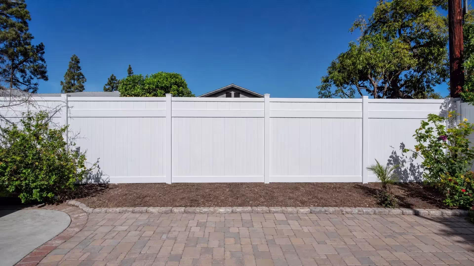 A multicolor paver patio borders this White vinyl privacy fence beside a stone edged raised soil bed