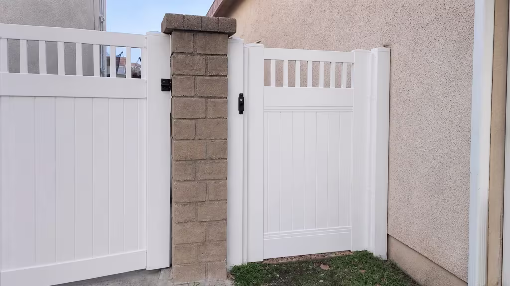 In a narrow passage between two stucco walls, a White vinyl gate carries its vertical picket top above solid privacy boards